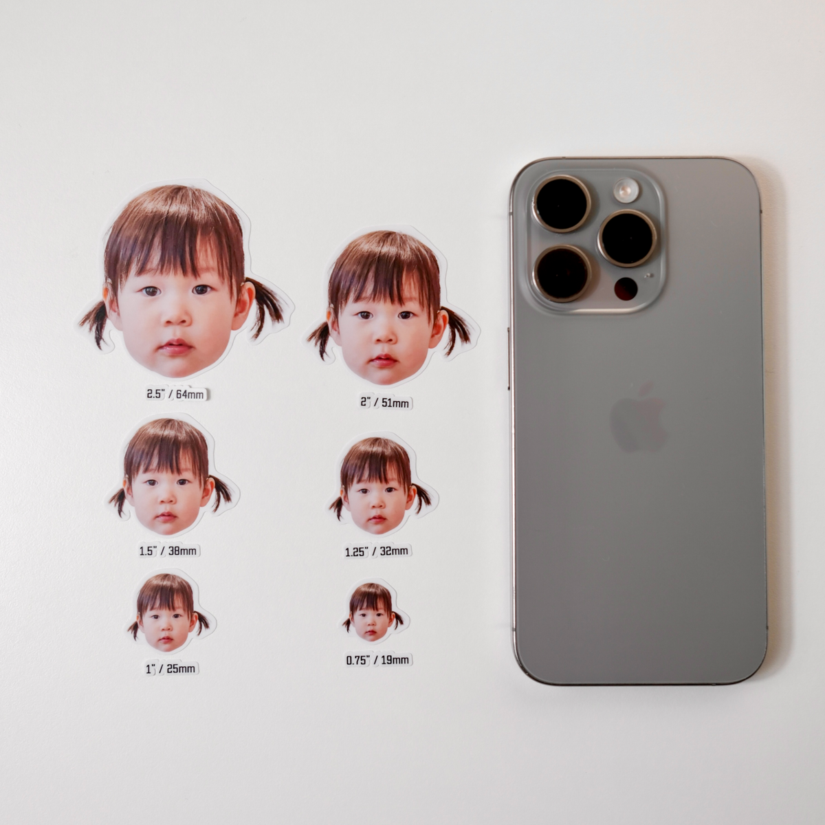 All six face sticker sizes, 2.5 inch down to 0.75 inch, laid out and labelled beside an iPhone for scale