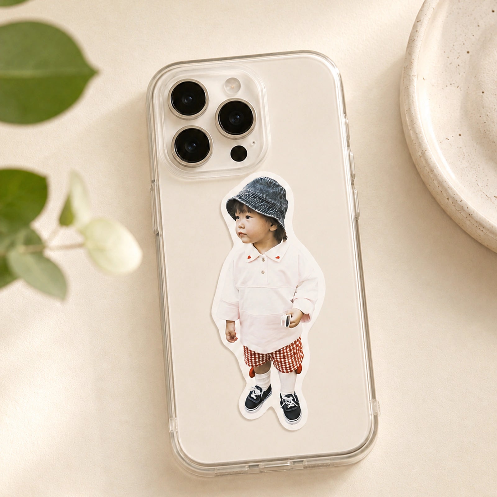 A die-cut full-body photo sticker of a toddler applied to a clear phone case, resting on an ivory surface beside eucalyptus leaves