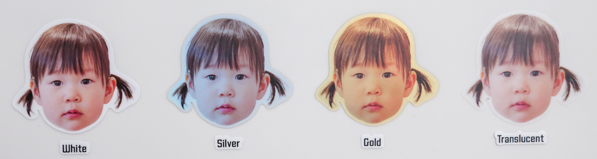 The same toddler face sticker printed on all four materials side by side, labelled White, Silver, Gold and Translucent