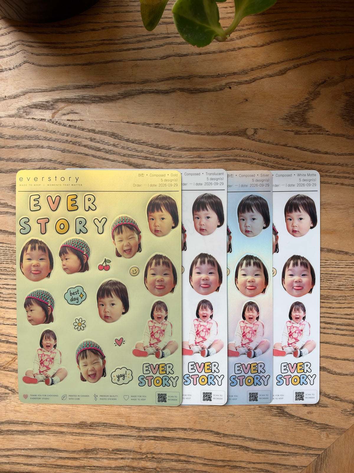 Four sticker sheets overlapping on a wooden table. All four together, left to right: Gold, Translucent, Silver, White Matte.